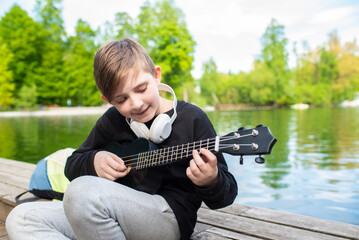 a little boy sits by the fountain, plays the black ukulele on a sunny day in the park