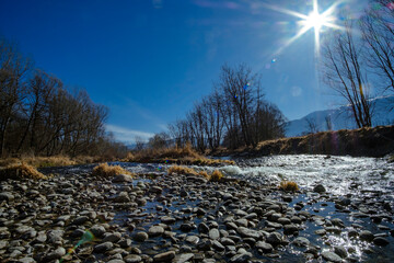 Cobble stones rolling on a wide river landscape on a winter leafless forest under a blue sunny sky with sun shining flare