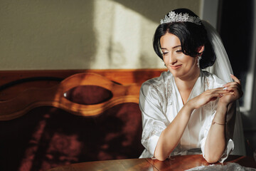 Portrait of a brunette bride sitting in a petticoat and touching her face. The silver crown is decorated with steel flowers. Gorgeous make-up and hair. Voluminous veil. Wedding photo. Beautiful bride