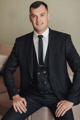 A man in a white shirt, black tie and black suit poses while sitting on a sofa. Front view. A stylish watch. Men's style. Fashion. Business