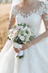 Cropped photo. The bride in a voluminous white dress and a long veil stands on the background of a field with a bouquet of white roses. Portrait of the bride. Beautiful makeup and hair.