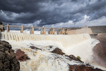 A dam with its flood gates open and a large volume of water flowing over rocks with storm clouds.
