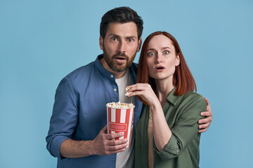 Excited middle aged couple of friends watching movie eating popcorn, looking at camera isolated on blue background