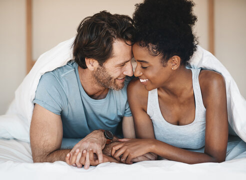 Happy interracial couple, bed and intimate morning in relax or bonding relationship at home. Man and woman smiling in joyful happiness for love, affection or relaxing weekend together in the bedroom