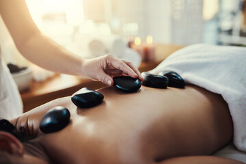 Woman, hands and rocks for back massage at spa in beauty relaxation or skincare on bed. Hand of masseuse applying hot rock or stones on female for physical therapy, zen or skin treatment at resort