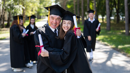 A group of graduates in robes outdoors. An elderly man and a young woman congratulate each other on...