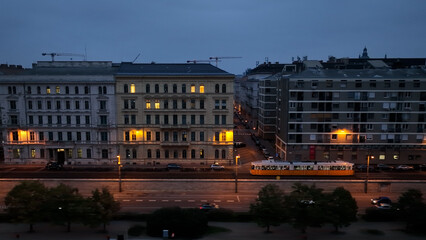 Fototapeta premium Aerial view of Typical Budapest Tram passing through city neighbourhood during blue hour, Aerial tracking follow shot wide angle, Hungary