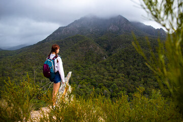 Naklejka na ściany i meble beautiful and brave girl standing at the top of the hill with large mountain in front of her; mighty mount barney in queensland, australia seen from yellow pinch lookout