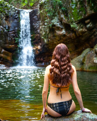 beautiful long hair woman in bikini sitting on the edge of a rock pool in front of large tropical waterfall at mount barney national park, queensland, australia