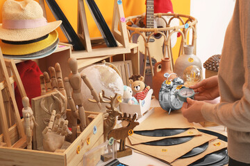Woman holding alarm clock near table with many different stuff indoors, closeup. Garage sale