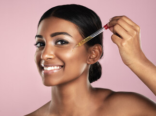 Fototapeta premium Face, skincare serum and smile of woman in studio isolated on a pink background. Portrait, cosmetics and Indian model with hyaluronic acid, essential oil or vitamin c dropper for healthy dermatology