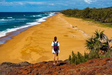 backpacker girl in white dress standing on the cliff admiring the panorama of beautiful beach with orange sand; pacific coast of australia in deepwater national park, queensland - obrazy, fototapety, plakaty