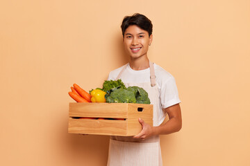 Happy greengrocer in T-shirt posing on background holding box full of fresh vegetables and fruits...
