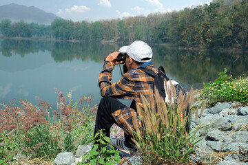 Asian boy in plaid shirt wears white cap, holding binoculars, standing on reservoir ridge during summer vacation and birdwatching activity, soft and selective focus, nature study and hobby concept.