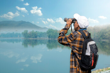 Obraz premium Asian boy in plaid shirt wears white cap, holding binoculars, standing on reservoir ridge during summer vacation and birdwatching activity, soft and selective focus, nature study and hobby concept.