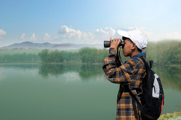 Asian boy in plaid shirt wears white cap, holding binoculars, standing on reservoir ridge during summer vacation and birdwatching activity, soft and selective focus, nature study and hobby concept.