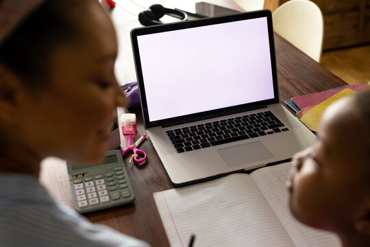 African american mother helping daughter with schoolwork using laptop at home, copy space on screen