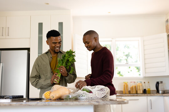 African american smiling gay couple removing groceries from mesh bag on kitchen island, copy space