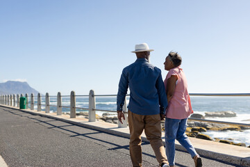Happy senior african american couple with sun hat walking on promenade at seaside