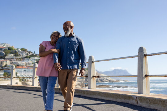 Happy senior african american couple walking and embracing on promenade at seaside