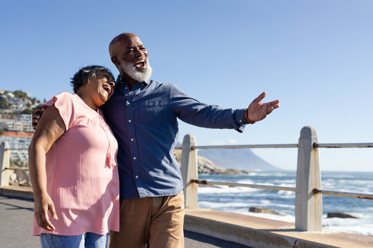 Happy senior african american couple walking and embracing on promenade at seaside