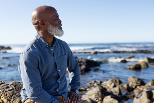 Thoughtful senior african american man sitting on rocks and admiring view at seaside