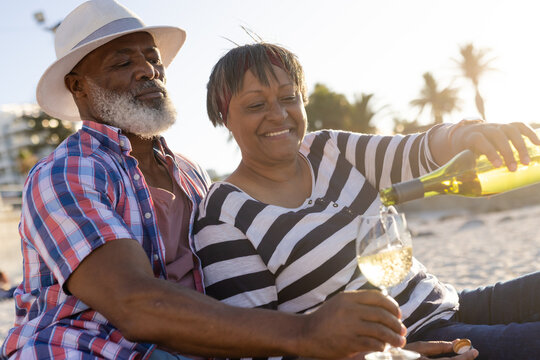 Happy senior african american couple having picnic and pouring wine at beach