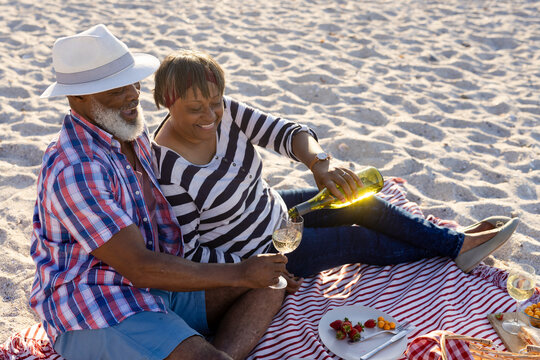 Happy senior african american couple having picnic and pouring wine at beach - Powered by Adobe