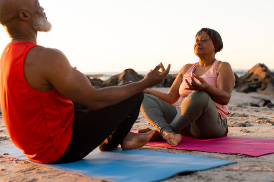 Happy senior african american couple practicing yoga on yoga mats and meditating at beach