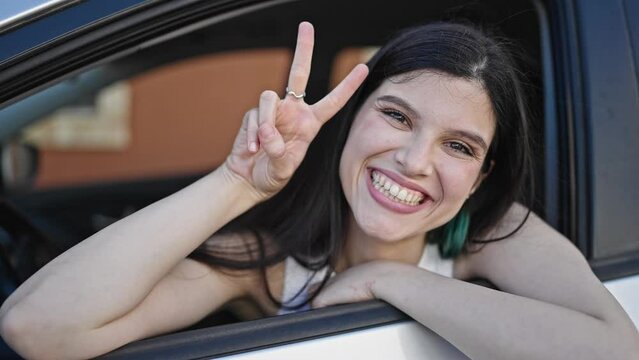 Young beautiful hispanic woman sitting on car doing victory gesture with fingers at street