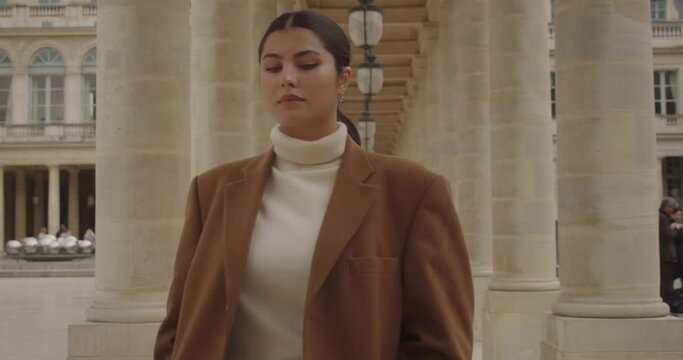 beautiful young woman with black hair walking in a public park with columns. Girl with coat in palais royal in paris in winter during a cold day.