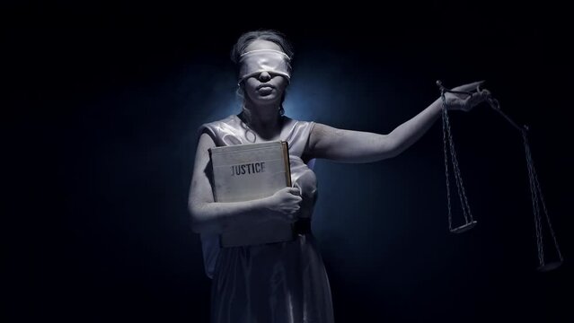 Portrait of a woman standing blindfolded, holding a book titled Justice, and raising a hand scale in a dark room. Image of the Themis as a concept of fair and unbiased justice