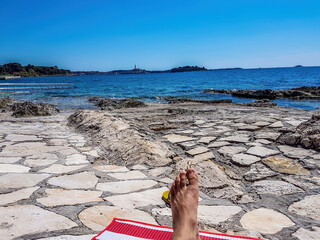 Relaxing on the beach. A human foot lying on a stony beach on a red towel. In the back there is bigger city with tall church tower. Clear and sunny day, perfect for suntanning.