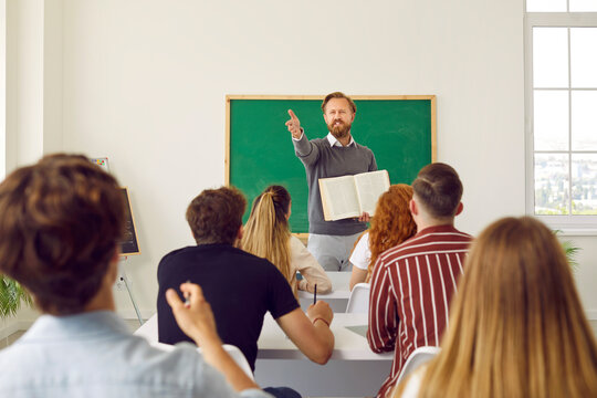 Middle aged professor lector showing on student in class for answer question. He standing near blackboard, students sitting backs to camera. Process of studying at highschool, college, university.