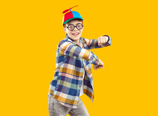 Portrait of happy funny preteen boy emotionally dancing. Cute joyful schoolboy wearing thick glasses, cap with propeller and casual clothing having fun over isolated yellow background © Studio Romantic