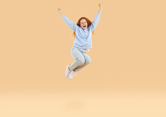 Fototapeta na wymiar Overjoyed excited teenage girl who feels euphoria joyfully shouts and jumps high with raised arms. Red-haired curly Caucasian girl in casual outfit jumping isolated on light beige background.