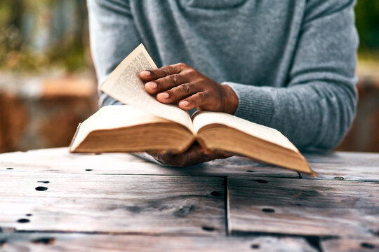 Hands, religion and a man reading the bible at a table outdoor in the park for faith or belief in god. Book, story and spiritual with a male christian sitting in the garden for learning or worship