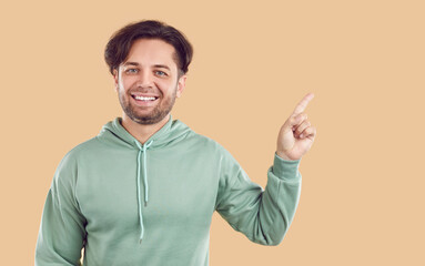 Smiling laughing happy bearded brunet man in mint hoodie is pointing index finger on copy space on beige background. Banner for advertisement, marketing, offer, sales. Sincere human emotions concept.