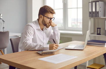 Focused businessman sitting at laptop computer and writing notes. Man accountant wearing glasses reviewing financial documents for analysis in office. Financial manager, bookkeeper, accountant,