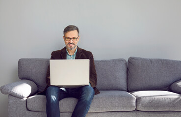 Successful mature businessman working on his laptop. Happy business man in smart casual clothes and eyeglasses using modern notebook computer while sitting on couch near grey wall at home or in office