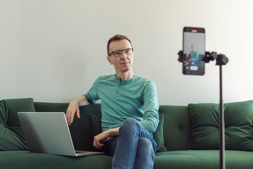 Handsome man, blogger, psychologist, sits at home on couch in front of phone camera and blogs,...