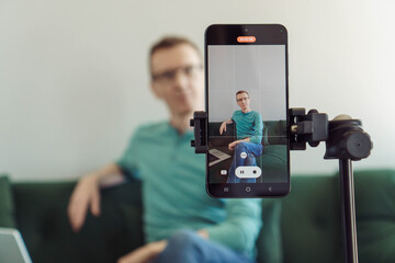 Unfocused man is sitting on sofa in background, focusing on phone on tripod with picture of camera shooting. Blogger. Modern technologies. Online training. Video recording of trainings. Video chat.