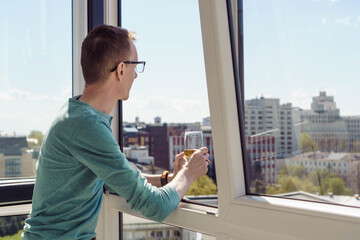 Thin man in turquoise T-shirt with long sleeves stands on balcony of house, apartment, office and looks at panorama of city below. City and buildings. Tourism and travel. Rest and relaxation.