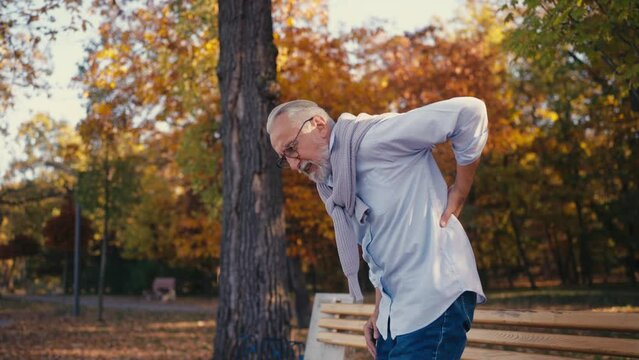 Senior man feeling acute back pain when standing up from a bench in a park