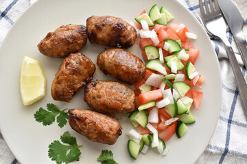 Sheftalia Cypriot lamb and pork Sausage. Grilled Sausages with Fresh Vegetables included sliced cucumber and sliced tomato on a white plate. Traditional Cypriot food.