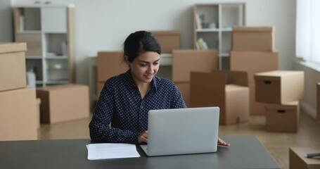 Indian businesswoman, small business owner using laptop, sit at desk, correspond to client or supplier of goods by e-mail working in warehouse, stacked carton boxes on background. SME owner workflow