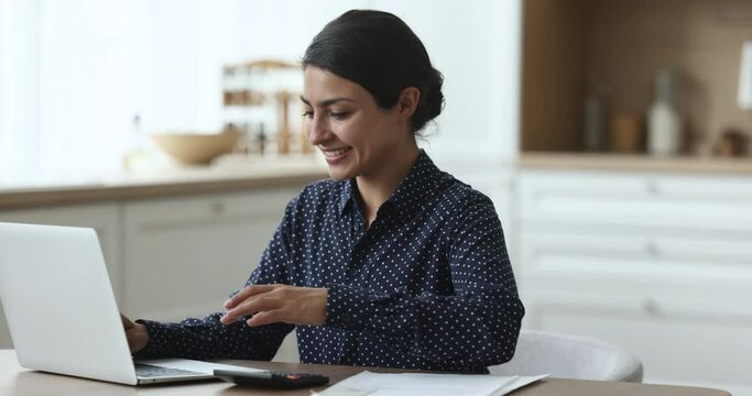 Smiling Indian woman sit at desk in kitchen use laptop and calculator, pay monthly bills through e-bank app, invoices, counting domestic budget, residential expenses, satisfied with monthly incomes