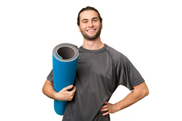 Young sport man going to yoga classes while holding a mat over isolated background posing with arms at hip and smiling