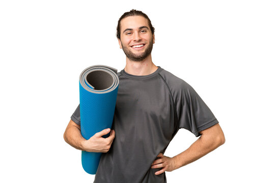 Young sport man going to yoga classes while holding a mat over isolated background posing with arms at hip and smiling