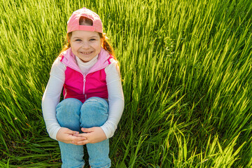 A cheerful girl sits in the thick green grass, clasping her legs with her hands, against the backdrop of the sunset. Walk outdoors in the city park